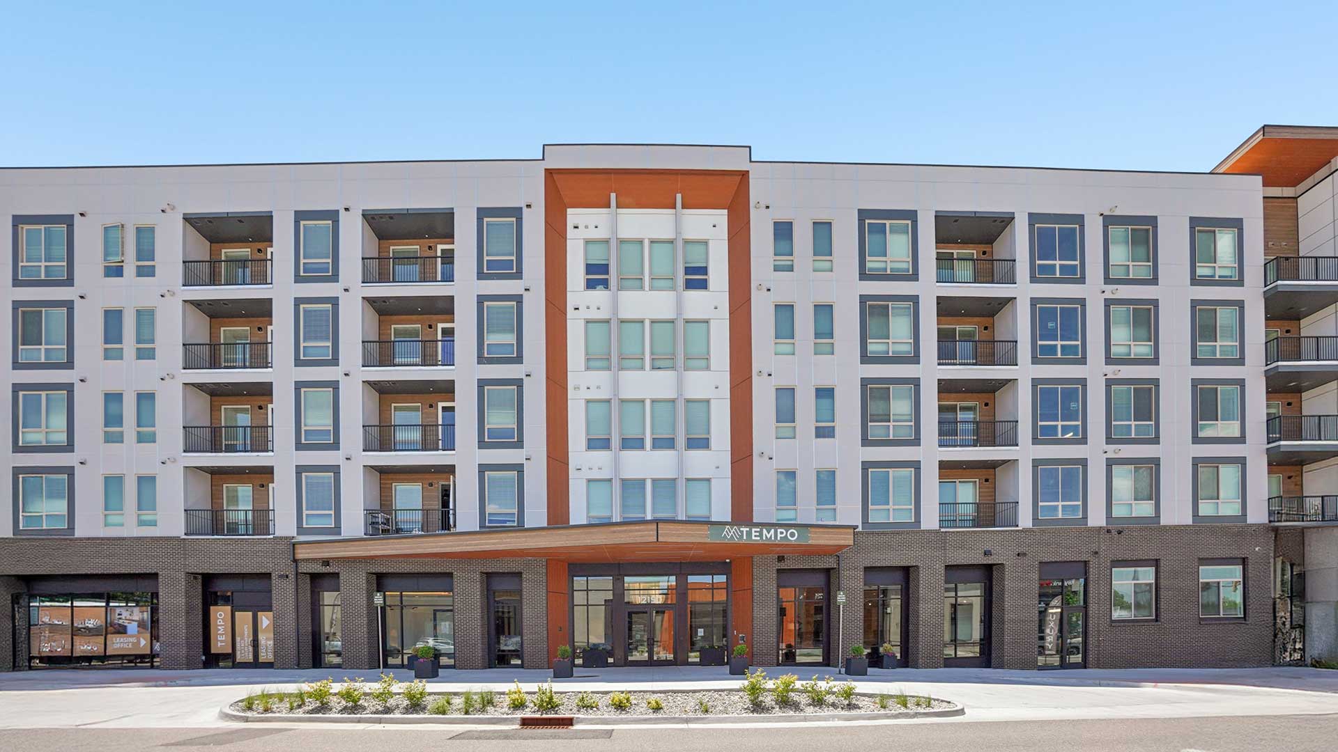 Front-facing exterior view of a modern multi-story apartment building with symmetrical architecture, balconies, large windows, retail-style ground floor spaces, landscaped entryway, and Tempo leasing entrance beneath a clear blue sky.