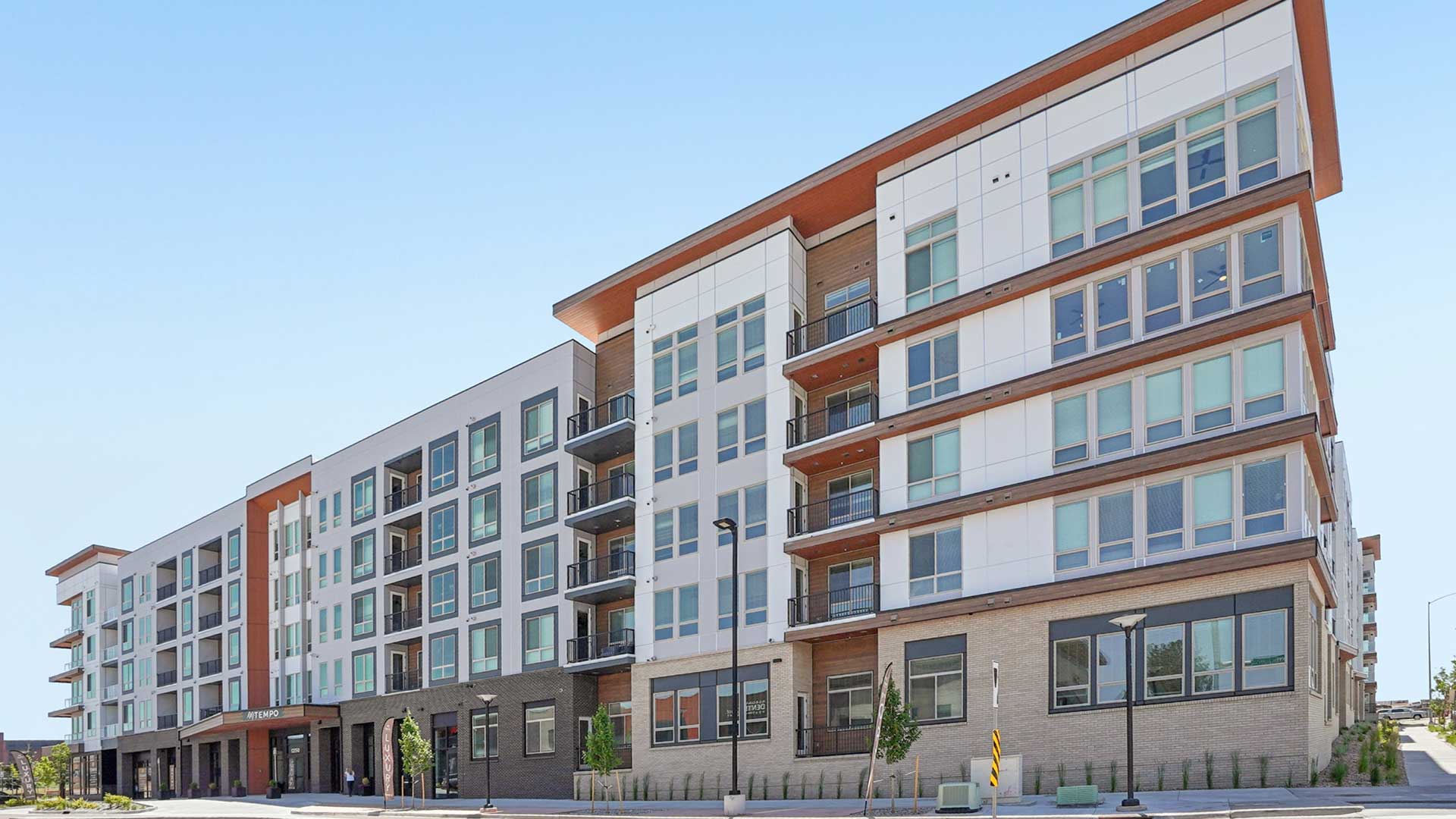 Exterior view of a modern multi-story apartment community with white and gray facade, wood accent panels, large windows, balconies, and street-level retail space under a clear blue sky.