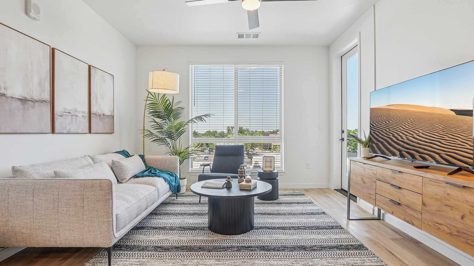 Modern apartment living room with a light-colored sofa, patterned area rug, coffee table, accent chair, large TV console, tall floor lamp, indoor plant, and balcony door with natural daylight streaming through large windows.