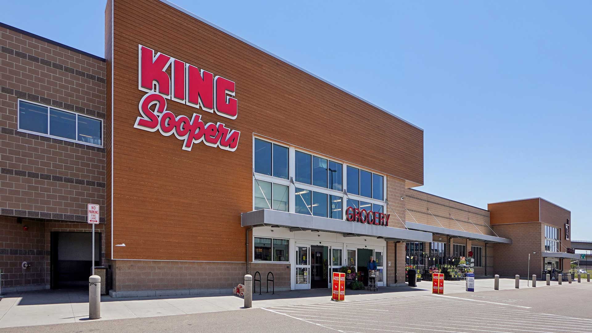 Exterior view of a modern King Soopers grocery store with large storefront signage, glass entrance doors, and a spacious sidewalk area under a clear blue sky.