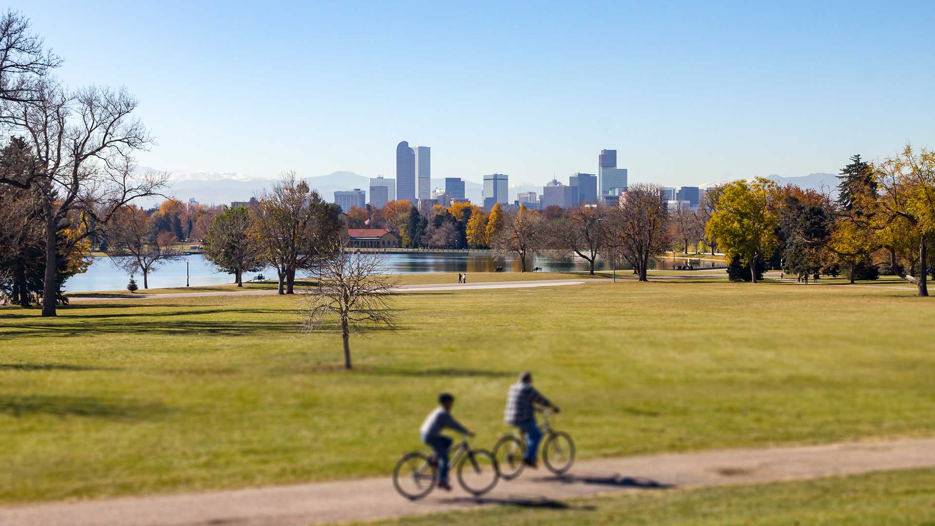 Cyclists riding through a large green park with a lake, trees, and the Denver skyline visible in the distance against the Rocky Mountains.