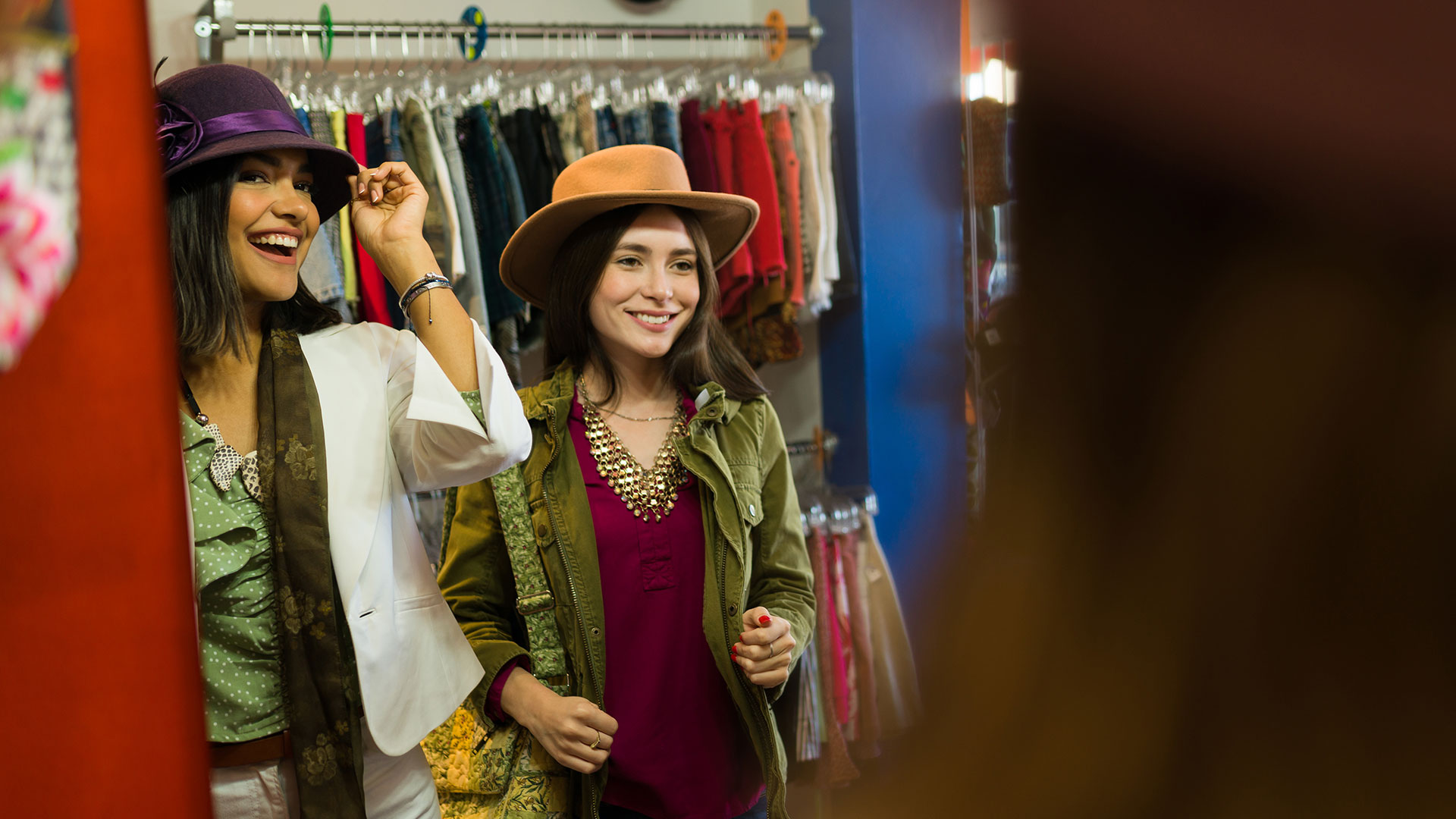 Two women shopping together in a boutique clothing store while trying on hats and browsing colorful apparel displays.