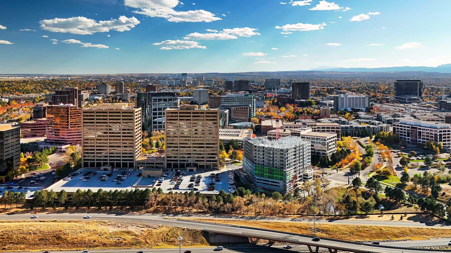 Aerial view of the Denver Tech Center featuring office towers, tree-lined streets, major roadways, and surrounding city development under a bright blue sky.