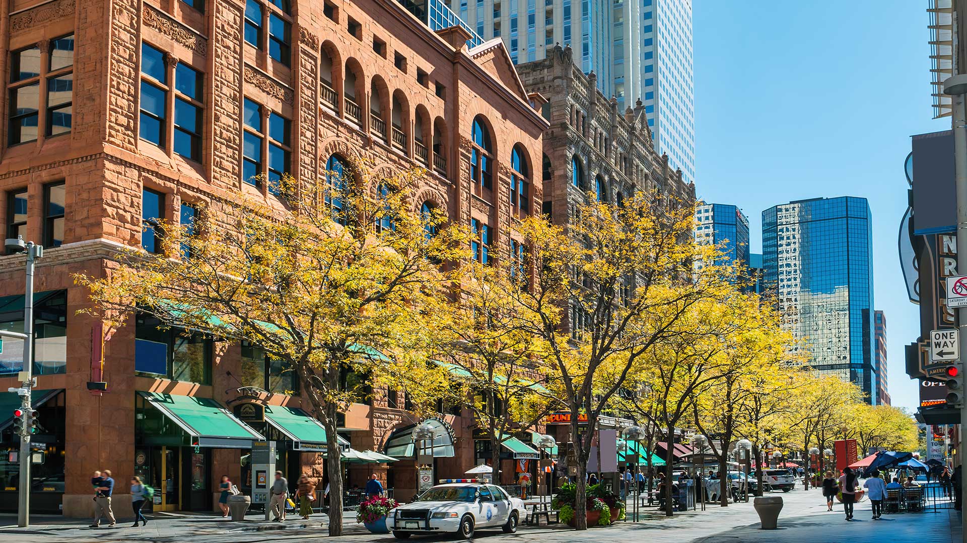 Tree-lined downtown Denver street with historic architecture, restaurants, patios, and pedestrian activity.