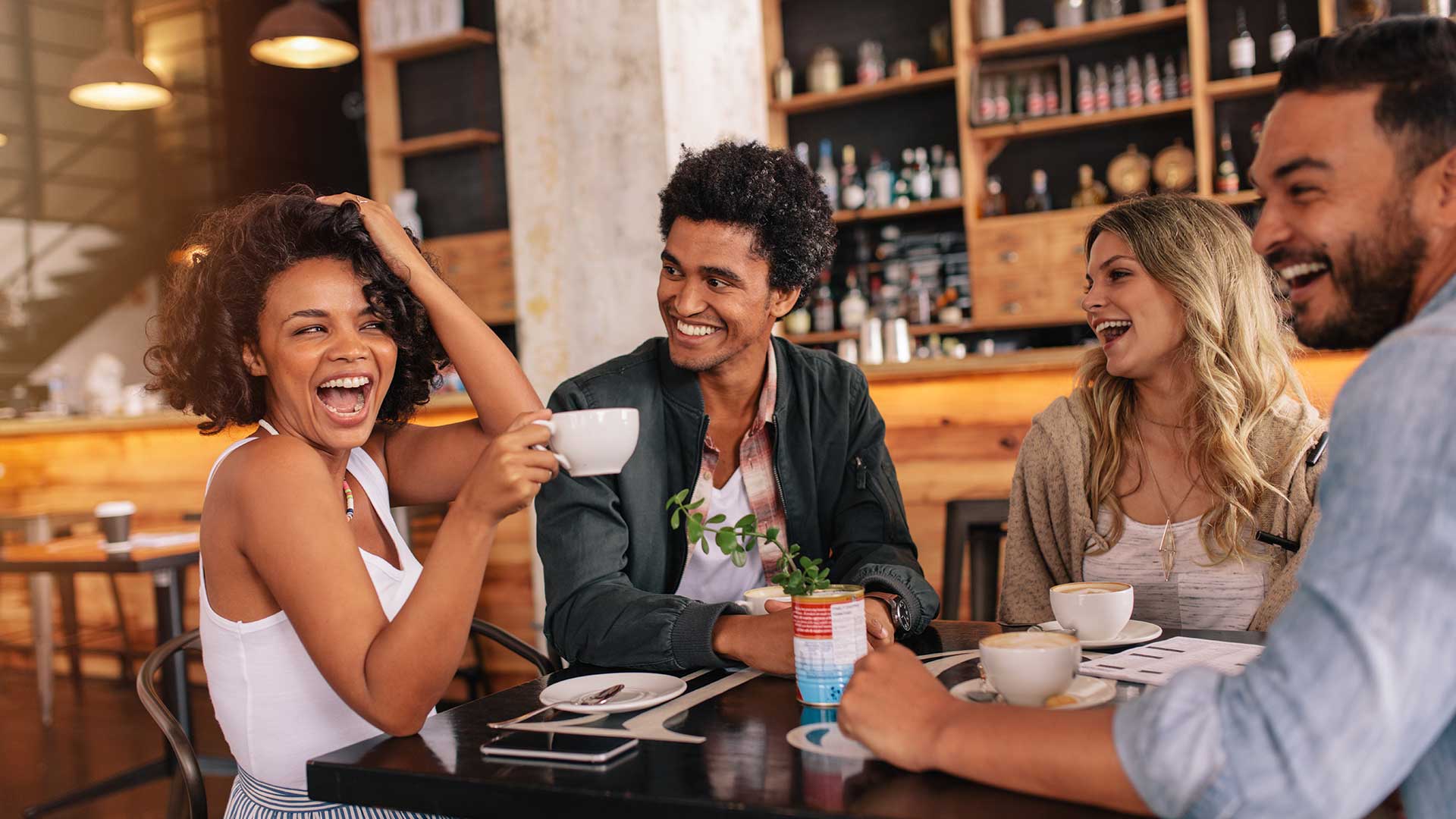 A group of friends laughing together at a modern coffee shop with warm lighting, wood accents, and coffee cups gathered around a table.